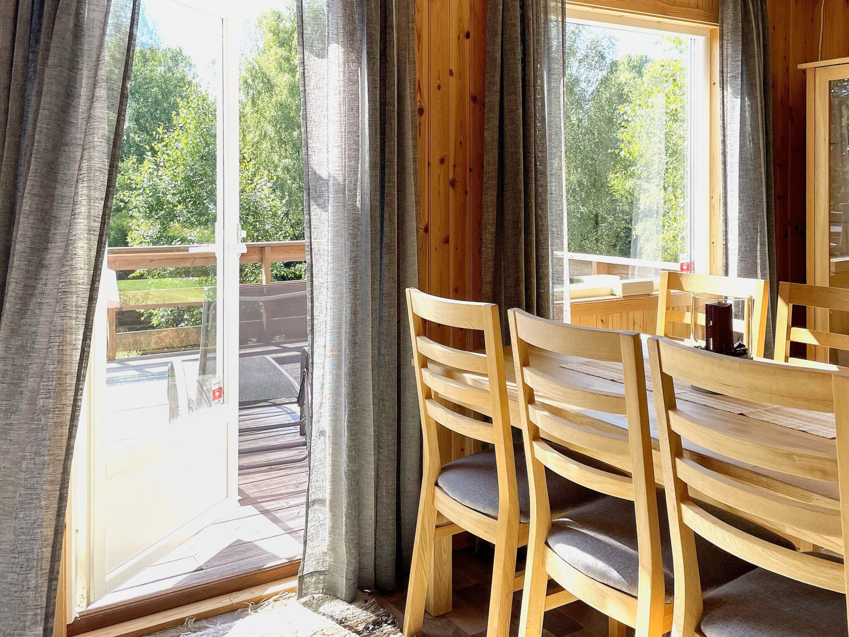 A wooden dining table with chairs, a window with curtains, and a view of a deck and greenery outside.