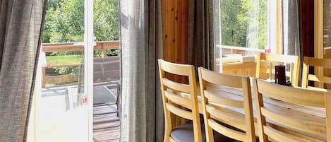Una mesa de comedor de madera con sillas, una ventana con cortinas y vistas a una terraza y follaje exterior.
