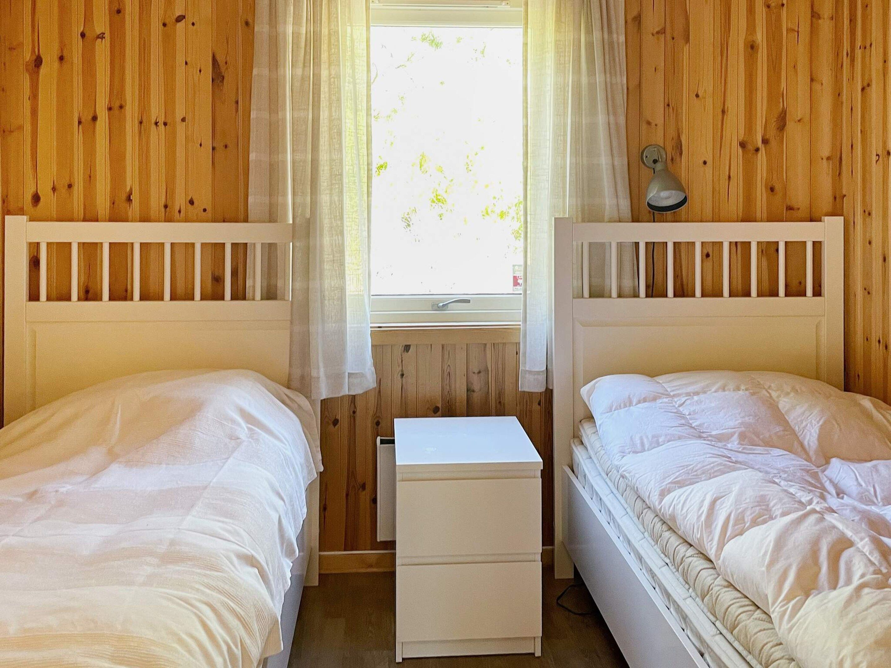 Quarto com duas camas, uma mesa de cabeceira pequena branca e uma janela com cortinas.