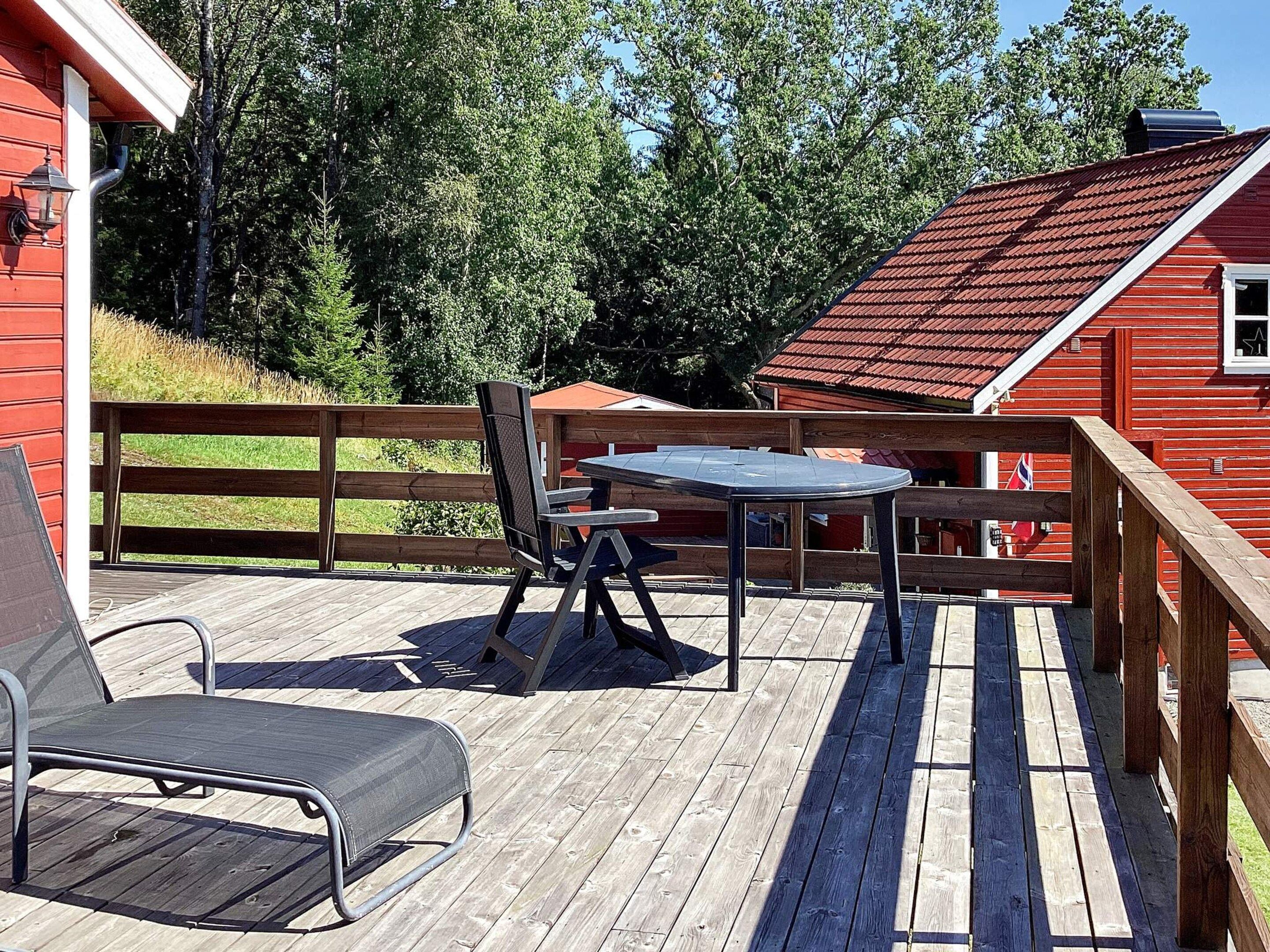 Una terraza de madera con una mesa, sillas y una tumbona, con un edificio rojo al fondo.