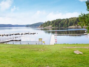 Blick auf einen See mit Steg, einer Rasenfläche und bewaldeten Hügeln.