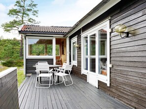 Eine Holzterrasse mit Tisch und Stühlen, großen Fenstern und Blick auf den umliegenden Wald.