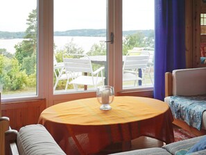 A room with a round table and chairs, a window with a view of a lake and trees, and a bed with a patterned bedspread. - 5 Person Holiday Home in Munkedal (Munkedal)