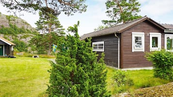 Uma cabana de madeira com telhado inclinado, rodeada por vegetação e um gramado.