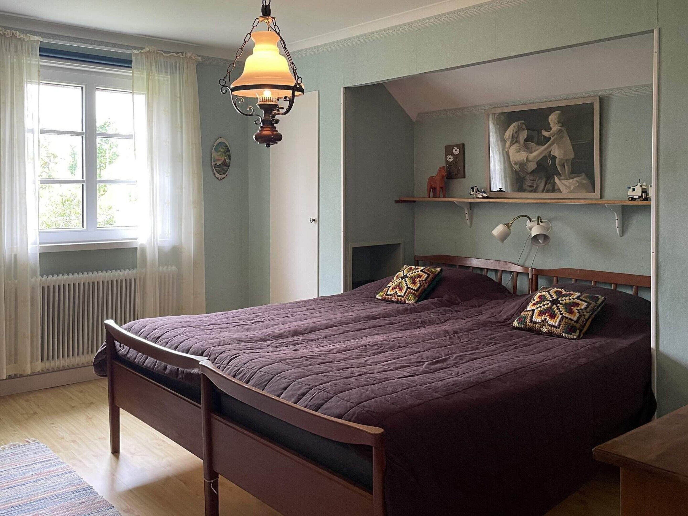 Un dormitorio con una cama grande, una lámpara colgante, un radiador, una ventana con cortinas y un cuadro en la pared.