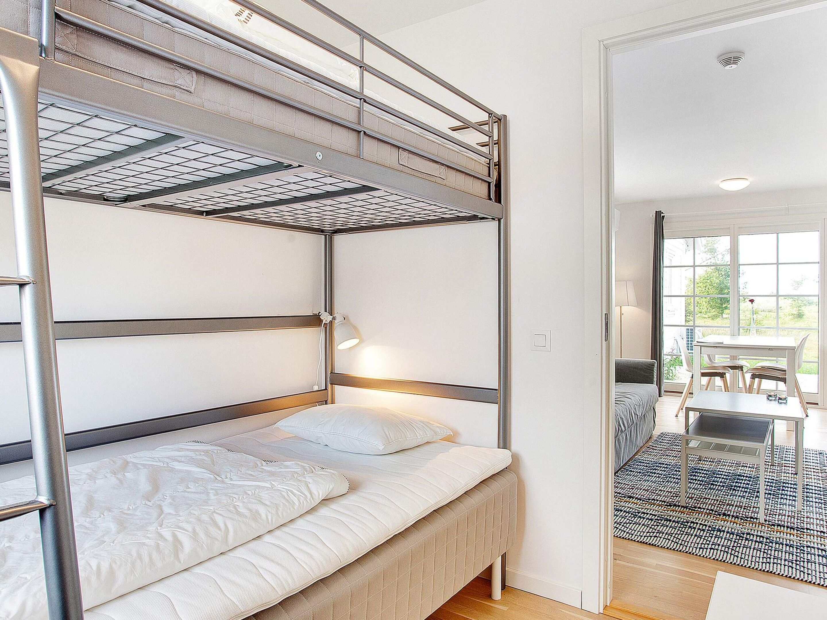 A bunk bed with a metal frame and white bedding, a bedside lamp, and a view of an outdoor dining area through a window.