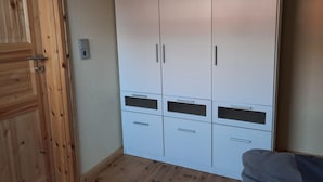 A room with a wooden door, a white wardrobe with multiple drawers and doors, and a bed with a grey blanket. - Ferienhaus am Vilzsee, Mirow (Mirow)