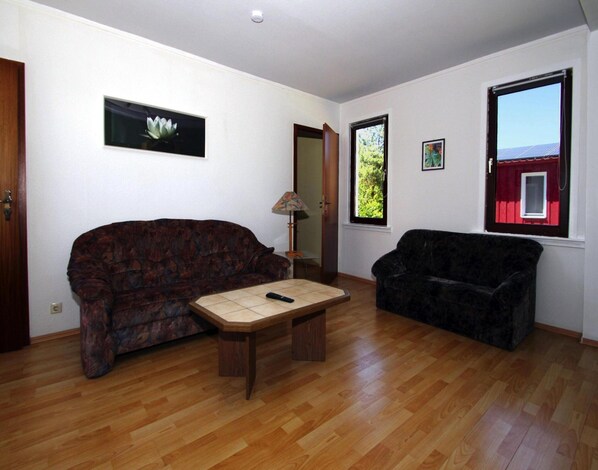 A living room with two sofas, a coffee table, and a lamp. There are windows with views of greenery and a red building. - Ferienhaus am Vilzsee, Mirow (Mirow)