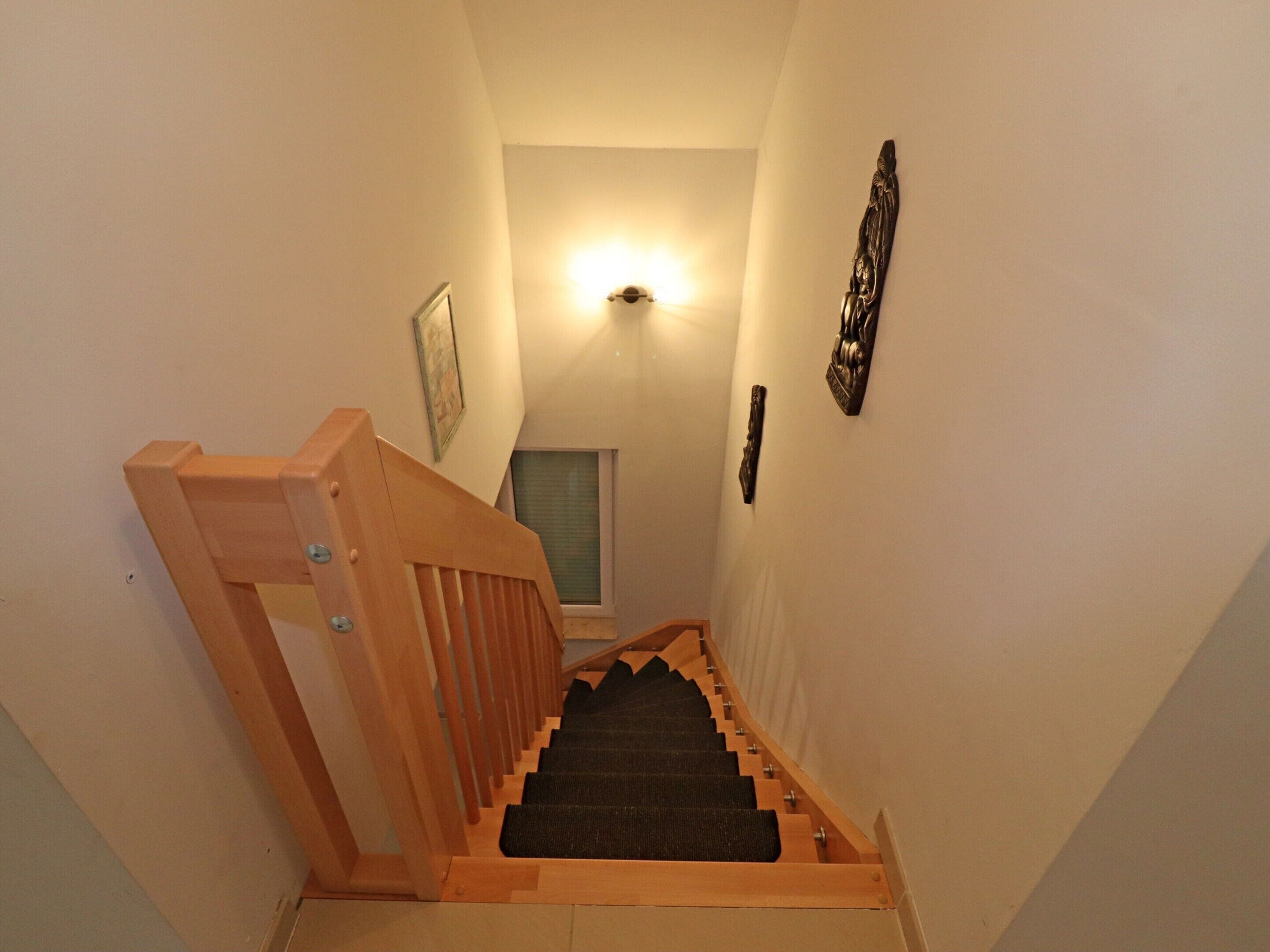 Een smalle trap met houten leuningen en een tapijtvloer, die uitkomt op een plateau met een lamp en een lijstwerk met een afbeelding.