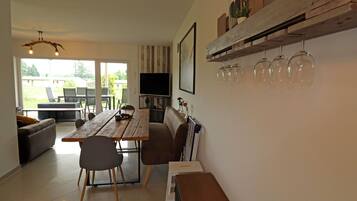 A dining area with a wooden table, chairs, and a television. There is a sofa and a bookshelf with decorative items.