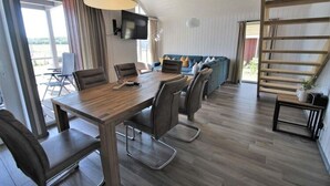 Ein modernes Wohnzimmer mit einem Holzesstisch, bequemen Sitzgelegenheiten, einem Fernseher und Blick ins Freie.