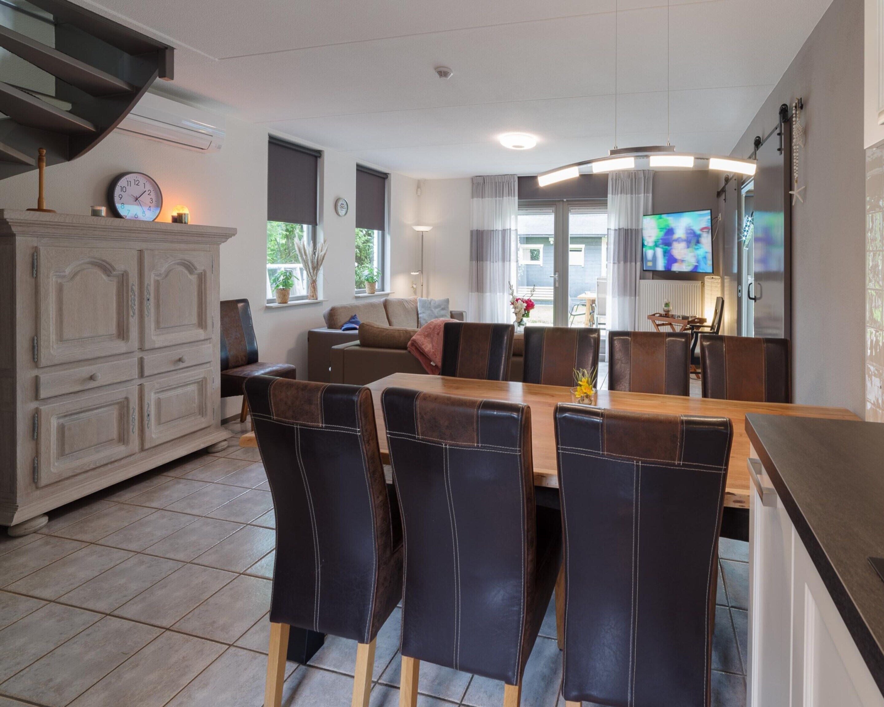 Une cuisine moderne avec un coin repas, comprenant une grande table en bois, des chaises en cuir foncé, un four encastré et un téléviseur.
