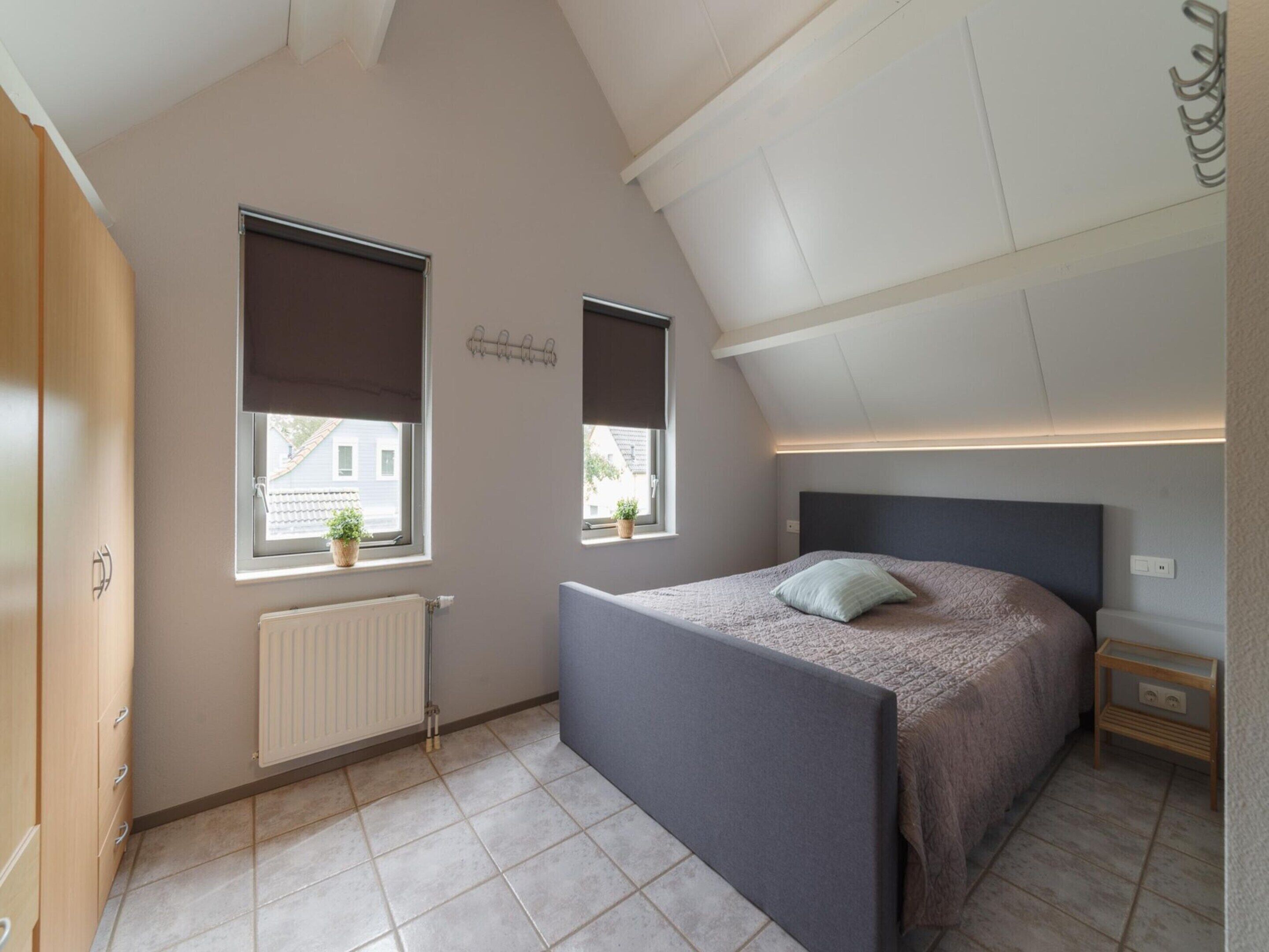 Une chambre avec un plafond en pente, un lit avec une tête de lit, deux fenêtres équipées de stores, un radiateur et une petite table de chevet.