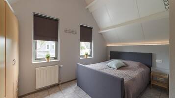 Une chambre avec un plafond en pente, un lit avec une tête de lit, deux fenêtres équipées de stores, un radiateur et une petite table de chevet.