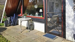 A patio with a table and chairs, a potted plant, and a window with a view of a house and trees. - Holiday Home in Burhave Near the sea (Butjadingen)