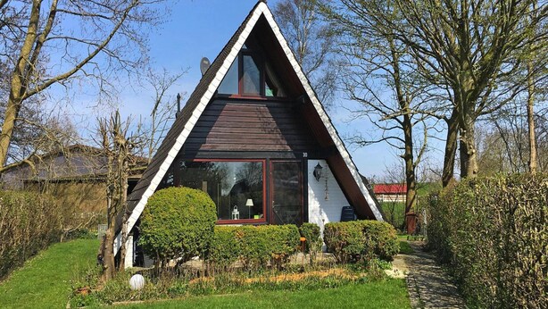 A modern, triangular-shaped house with a large glass window, surrounded by well-maintained gardens and trees. - Holiday Home in Burhave Near the sea (Butjadingen)