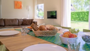 En frukostuppdukning med ett träbord, tallrikar, glas, en korg med bröd och skålar med frukt.
