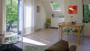 Ett ljust och luftigt vardagsrum med öppen planlösning, ett matområde, en soffa, ett glasbord och utsikt över en trädgård genom stora fönster.