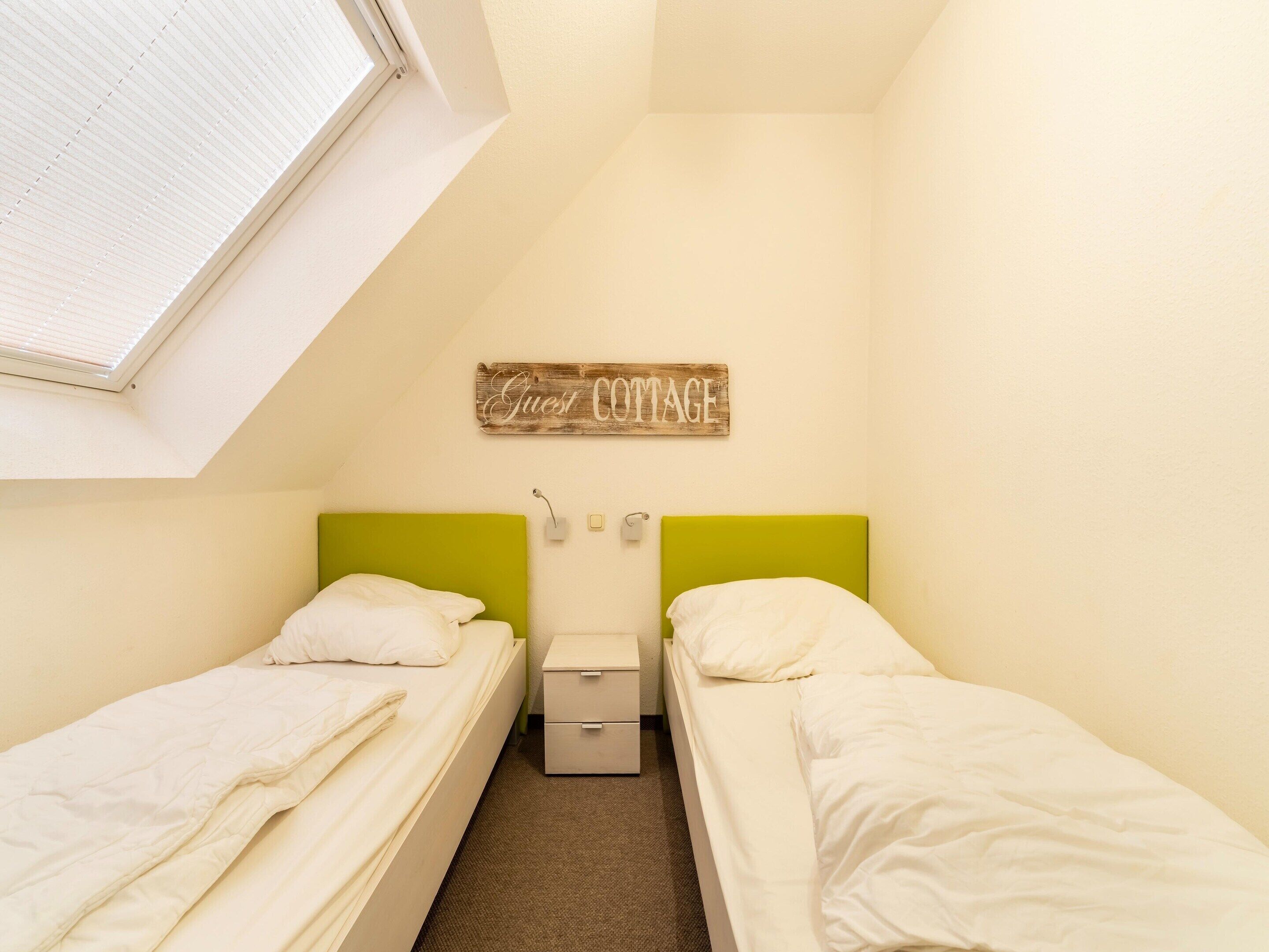 A small, clean room with two single beds, a small bedside table, a wall-mounted light, and a window with blinds.