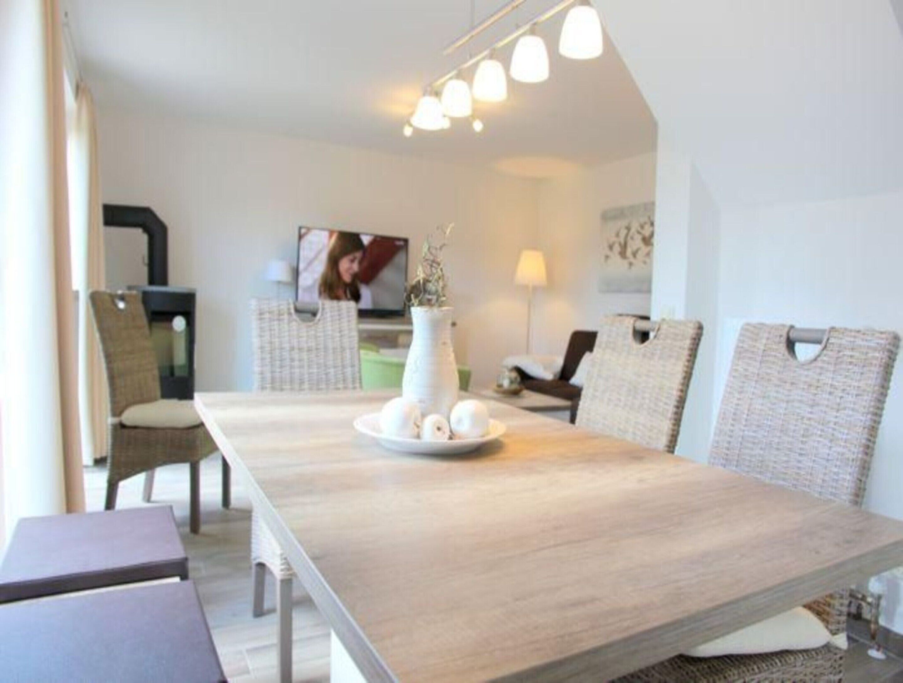 A modern dining area with a long wooden table, wicker chairs, and a television in the background.