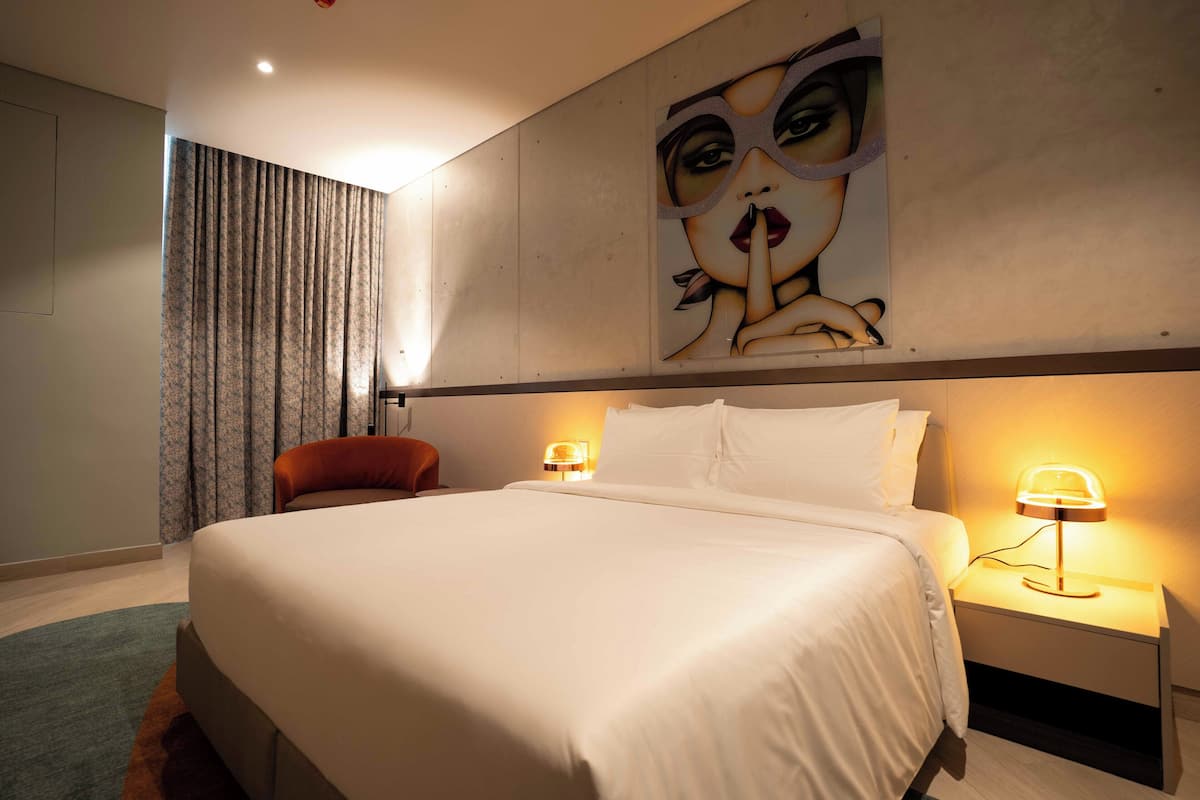 A hotel room with a large bed, two bedside lamps, a red chair, a painting on the wall, and a curtain.