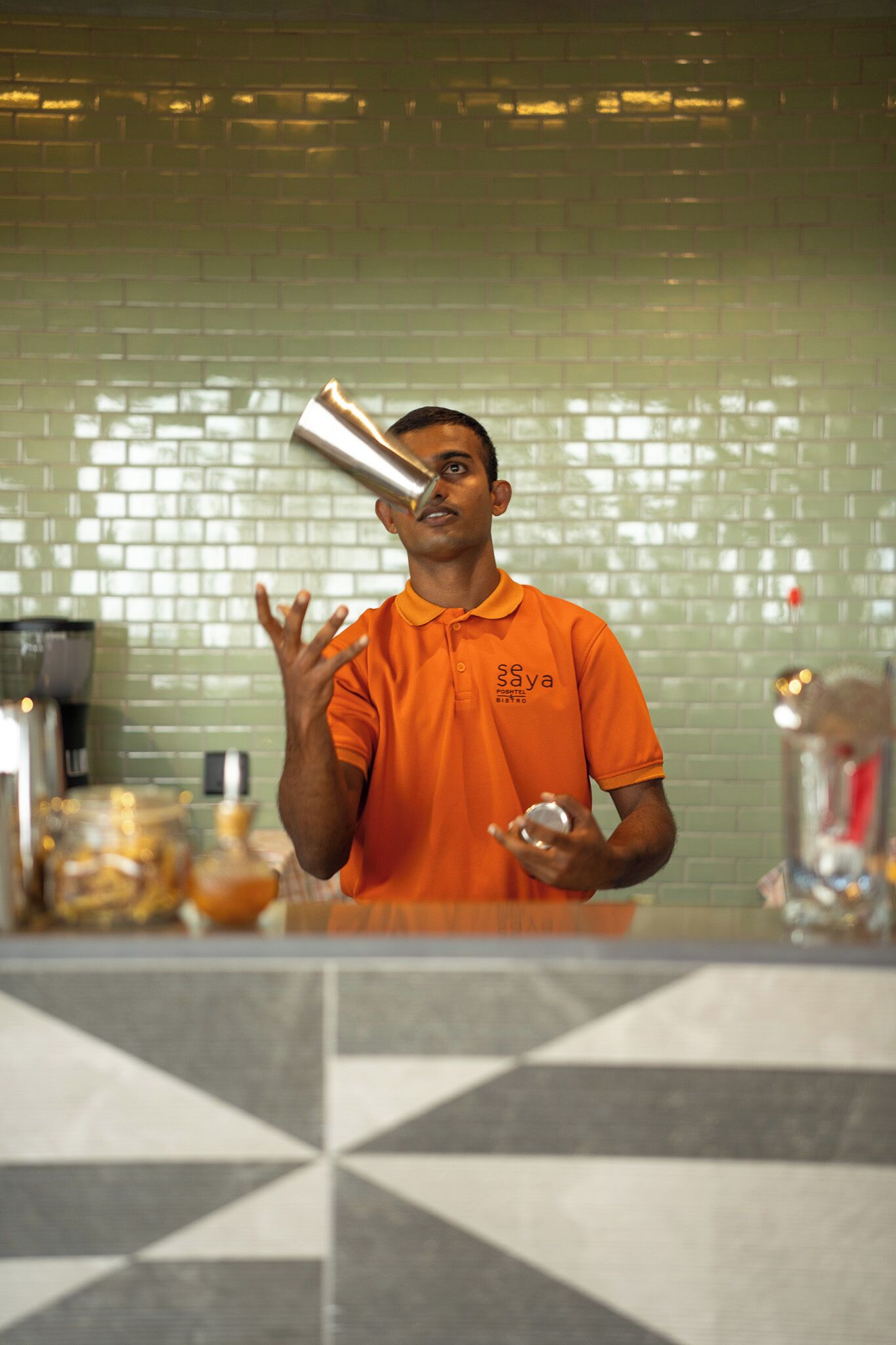 A person in an orange shirt with a logo, holding a drink and a bottle, standing in a kitchen with a tiled backsplash.
