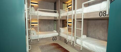 A dormitory room with bunk beds numbered 03 to 08, a wooden floor, and a patterned rug.