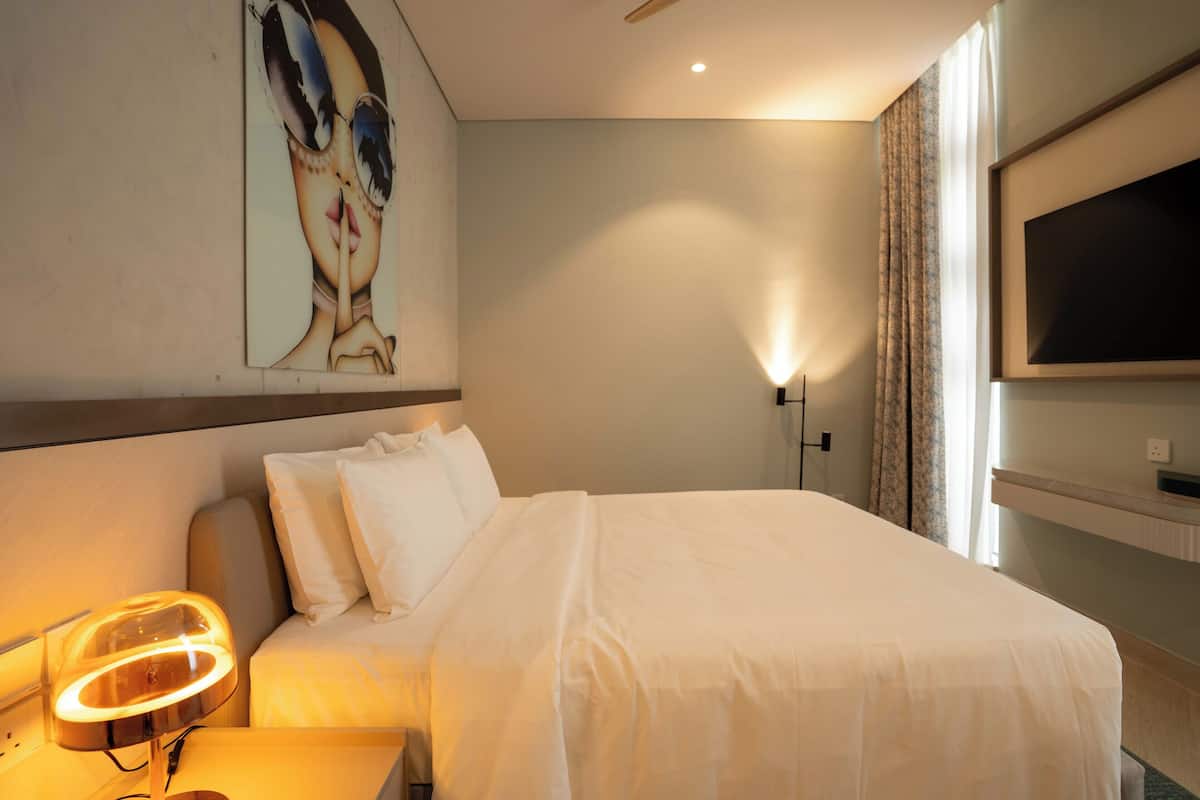 A hotel room with a bed, a bedside lamp, a TV mounted on the wall, and a painting on the wall.