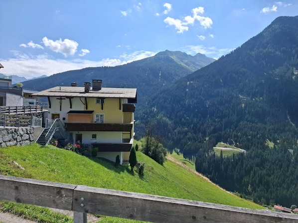 A multi-story building with a staircase leading to the entrance, surrounded by lush green hills and mountains. - Holiday Apartment in Kappl Near ski Resort (Kappl)