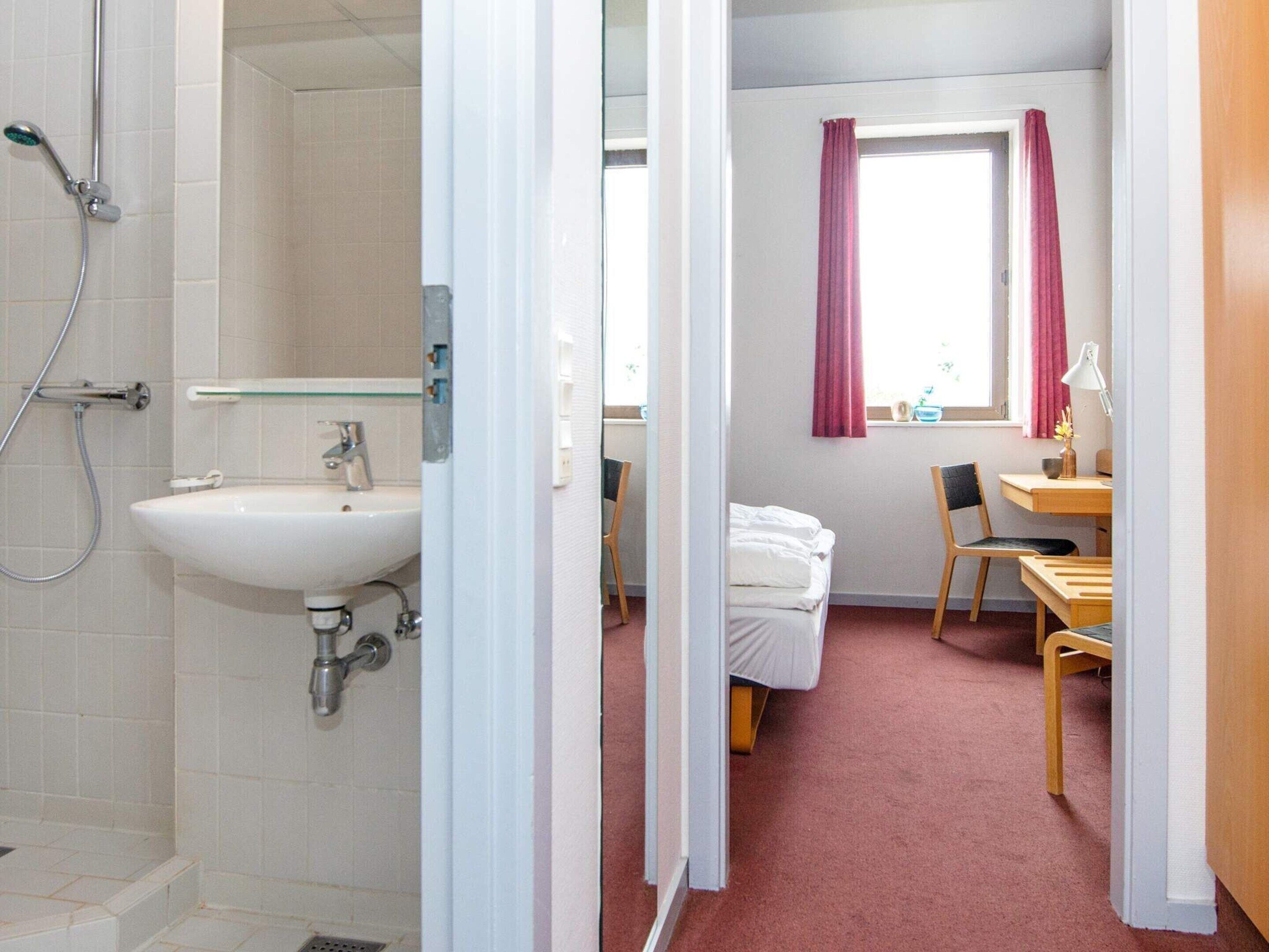Une chambre d’hôtel équipée d’une douche, d’un lavabo, d’un lit, d’un bureau, d’une chaise et d’une fenêtre avec des rideaux.