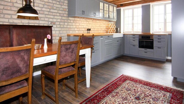 Une cuisine avec des poutres en bois, des meubles de couleur grise, un mur en briques et un coin repas comprenant une table et des chaises.