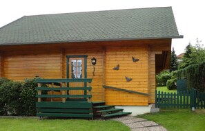 Une cabane en bois avec un toit vert, une véranda à l’avant dotée d’un banc et un petit escalier menant à l’entrée.