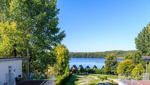 A view of a lakeside property with a wooden deck, a small building with a red roof, and a clear blue sky. - Elegant Eingerichtete Ferienwohnung (Mirow)