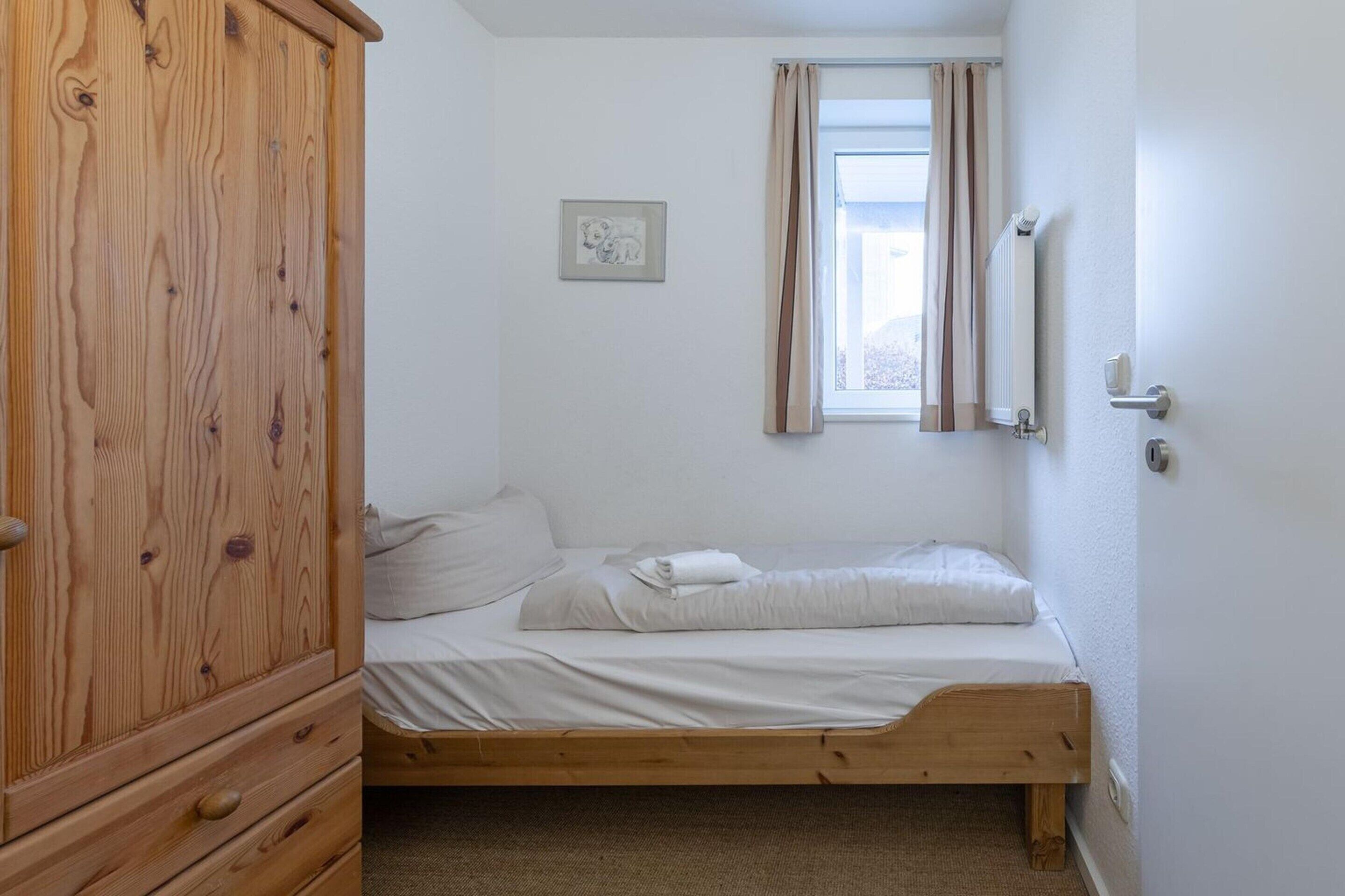 A small, single-bed room with a wooden wardrobe, a radiator, and a window with curtains.