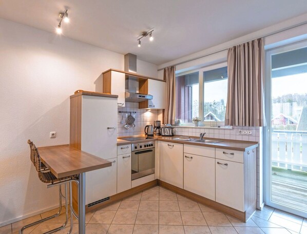 A compact kitchen with a built-in oven, a sink, and a window with curtains. - Appartement im Ferienpark in Rechlin (Rechlin)