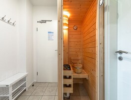 Una sauna pequeña y compacta con paredes de madera y un banco. Se observa una puerta blanca con manija y un ventanal pequeño. En la pared se ve un reloj.