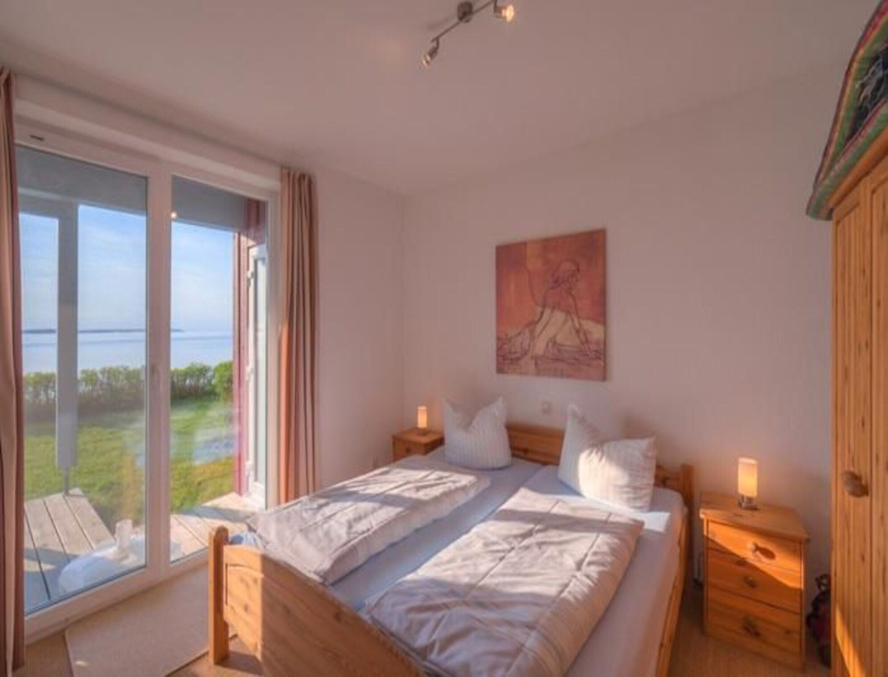 A bedroom with a large window, a bed, two bedside tables, and a painting on the wall.