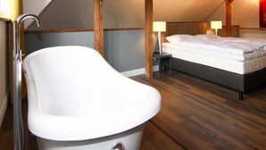 A bedroom with a sloped ceiling, wooden beams, a bathtub, a bed, and two bedside lamps. - Ferienhaus Birgit, Wendorf (Kuhlen-Wendorf)