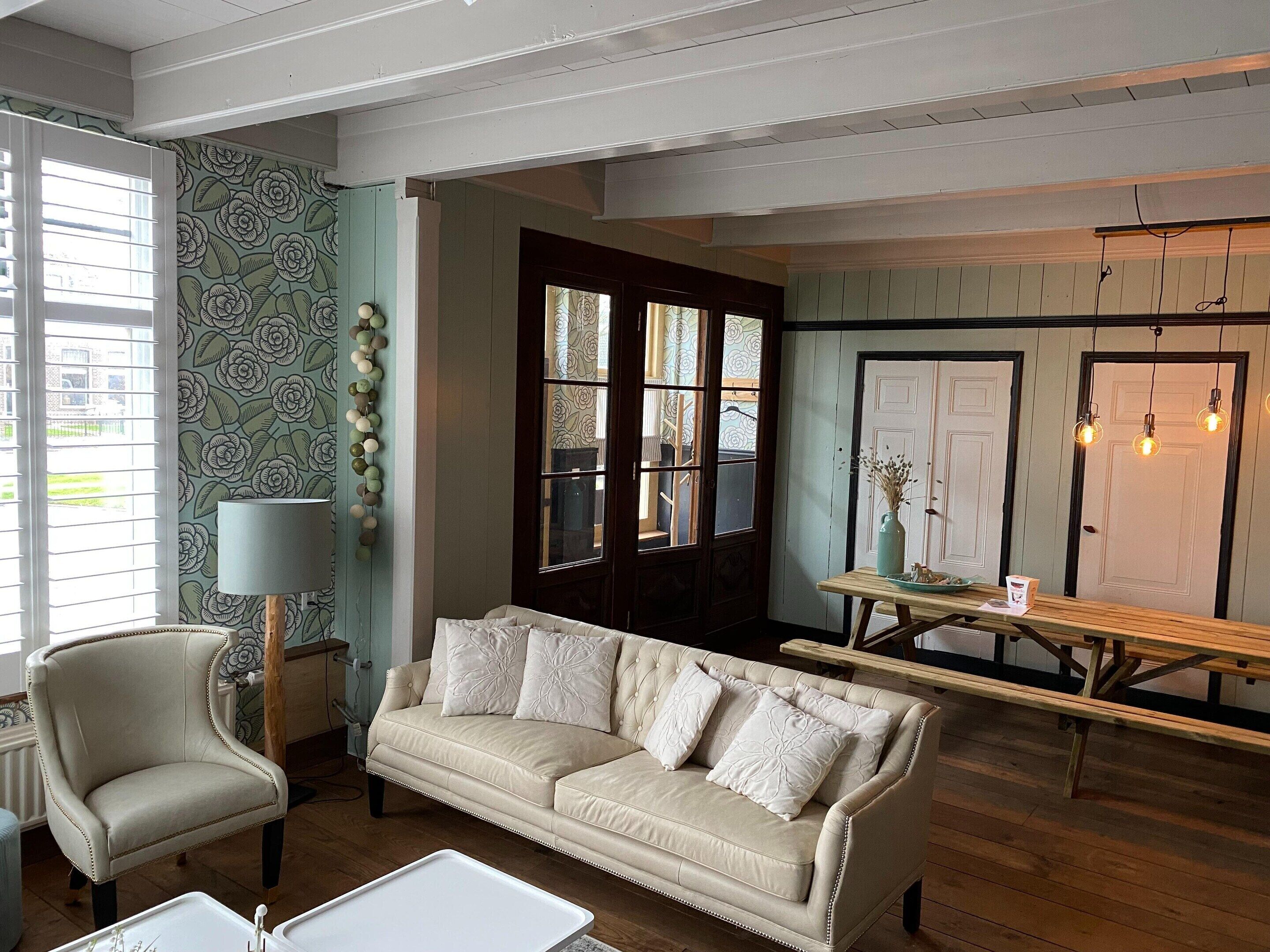 A cozy living room with a beige sofa, a wooden coffee table, and a lamp. There is a patterned wallpaper and a door with glass panels.