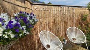 Zona de pati amb una tanca de bambú trenat, dues cadires blanques de vimetla i un cistell de flors morades i blanques.