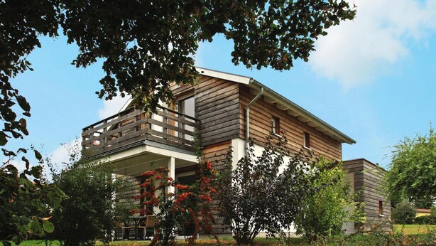 A two-story wooden house with a balcony, surrounded by trees and a garden.