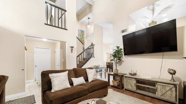 Ein Wohnzimmer mit einem braunen Sofa, einem Couchtisch mit Büchern, einem an der Wand befestigten Fernseher und einer Treppe im Hintergrund.