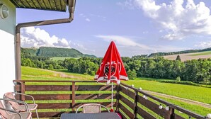 Une terrasse en bois avec des chaises et une table en osier, donnant sur un paysage verdoyant agrémenté d’un parasol rouge.