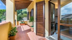 Un espacio al aire libre techado con suelo de baldosas, contraventanas de madera, plantas en macetas y una puerta de cristal que da a una habitación.