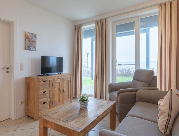 Un salon avec un meuble TV en bois, un canapé, une table basse et une vue sur l’extérieur grâce à de grandes fenêtres.