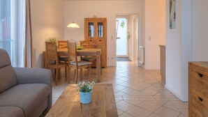 Ein Wohnzimmer mit einer Couch, einem Couchtisch mit Pflanze, einem Essbereich mit Tisch und Stühlen sowie einer Küchenzeile mit Schränken und einem Kühlschrank.