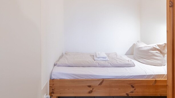 Een kleine kamer met een enkel bed, houten frame, wit beddengoed en een klein schilderijtje aan de muur.