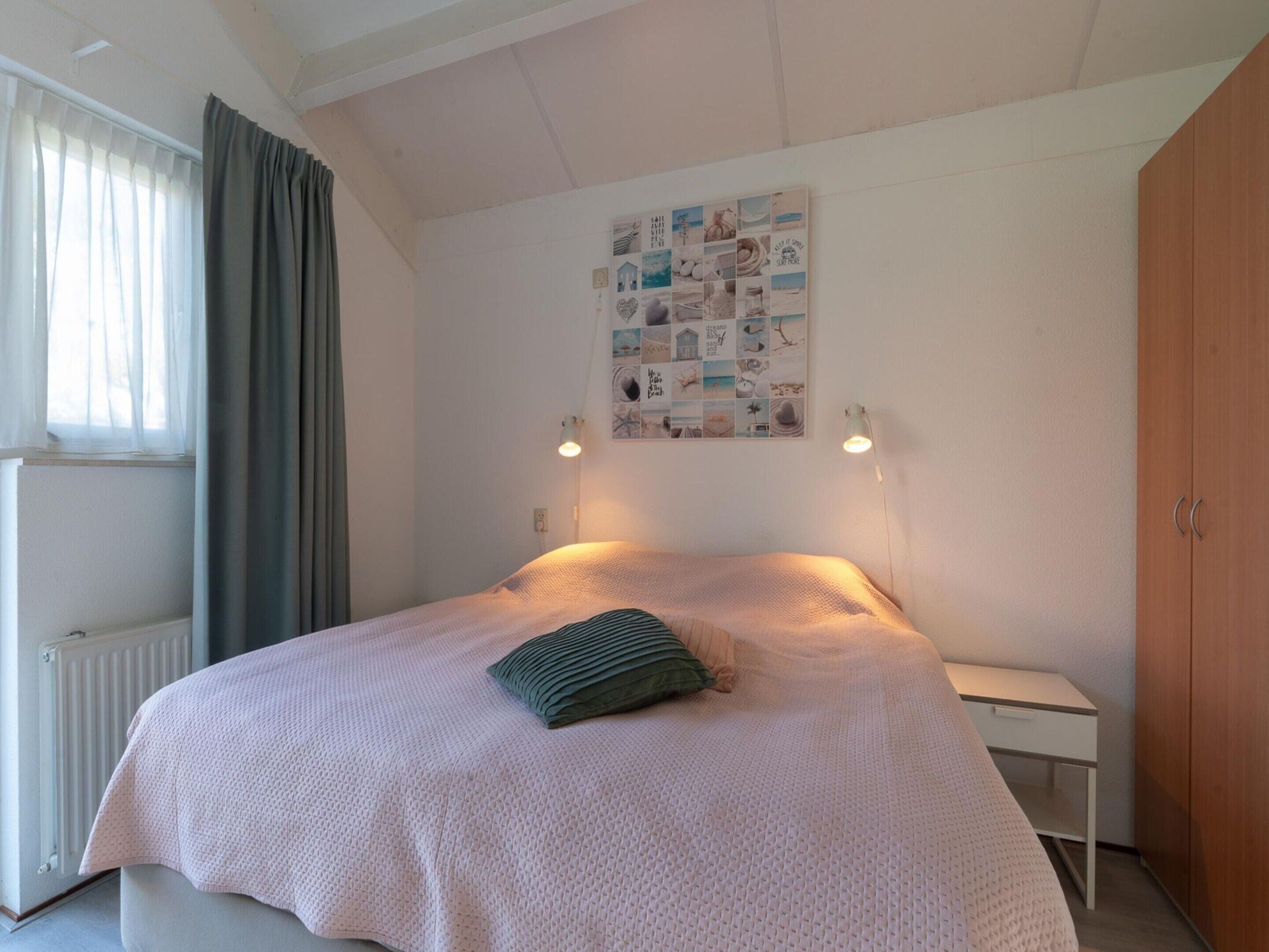 A bedroom with a bed, a bedside table, a wall-mounted light, a radiator, and a window with curtains.