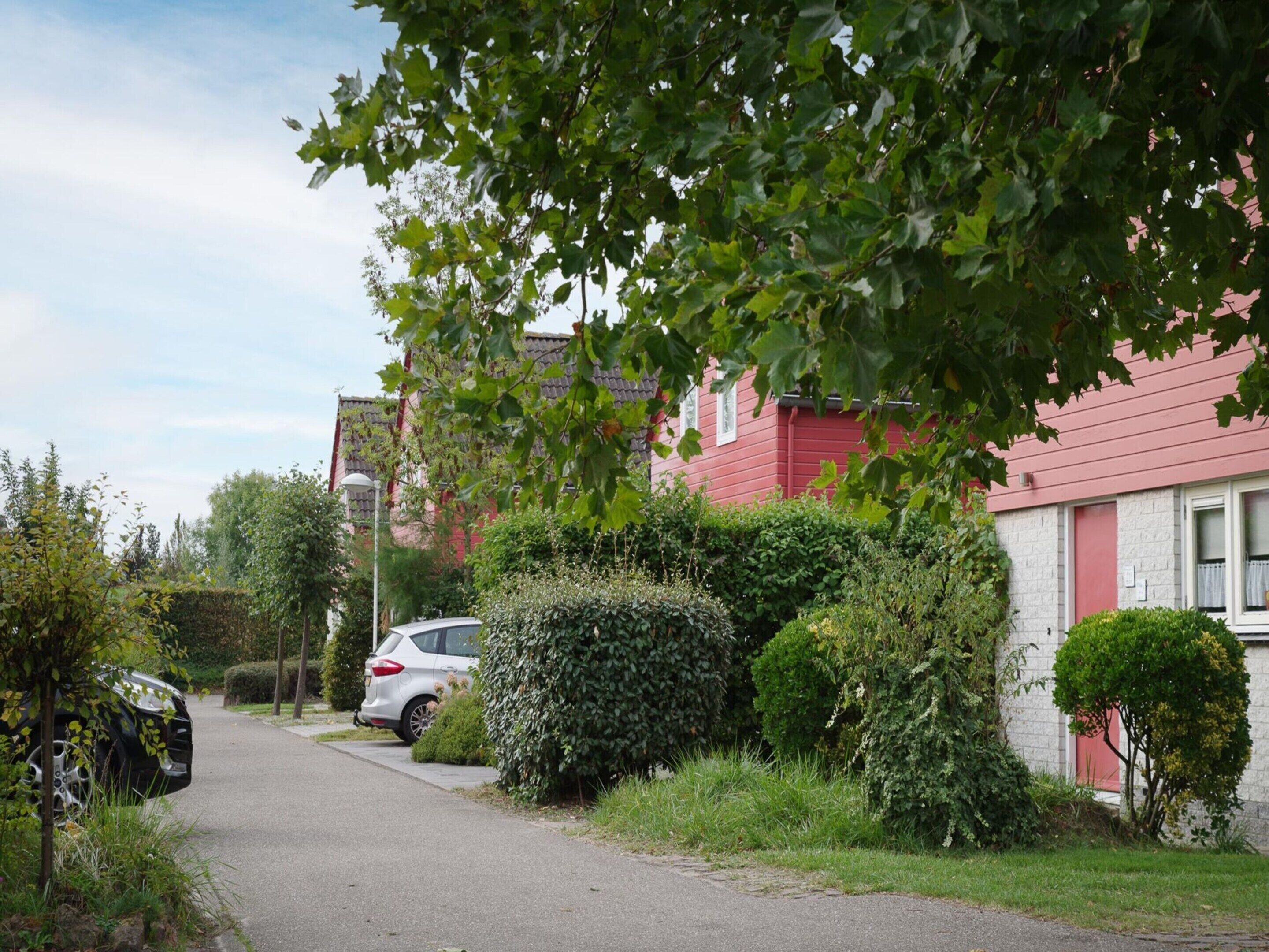 En bostadsgata med ett rött hus, en parkerad bil och en häck.