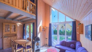 A wooden cabin interior with a dining area, a sofa, and a large window offering a view of the outdoors. - Ferienhaus im Skandinavischen Stil (Rechlin)